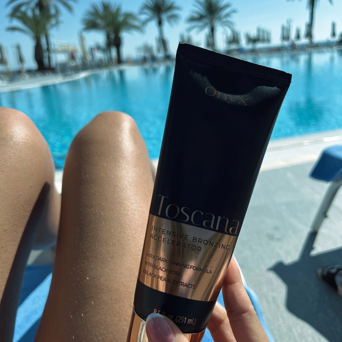 Loción bronceadora Toscana sostenida frente a piernas bronceadas junto a la piscina