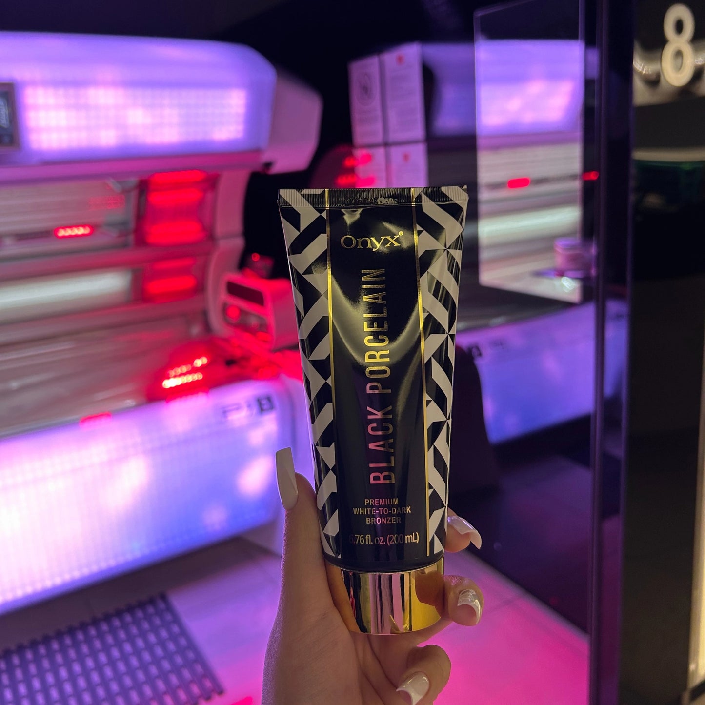 La loción bronceadora Onyx Black Porcelain sostenida frente a una moderna cabina de solarium por la noche, ambiente de producto lujoso