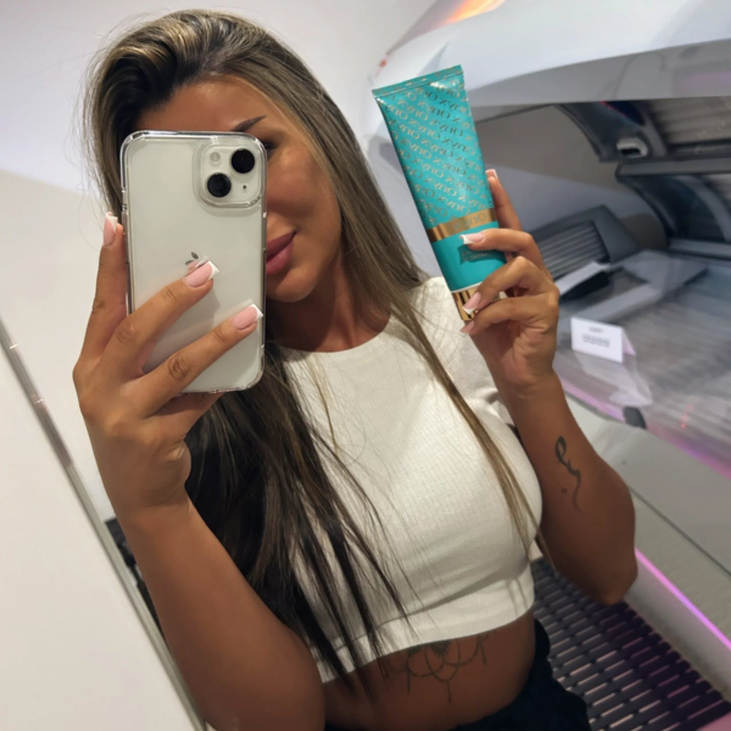 Mujer tomando una selfie en un salón de bronceado mientras sostiene la loción Onyx Dark Tanning Accelerator frente a una cama de bronceado.
