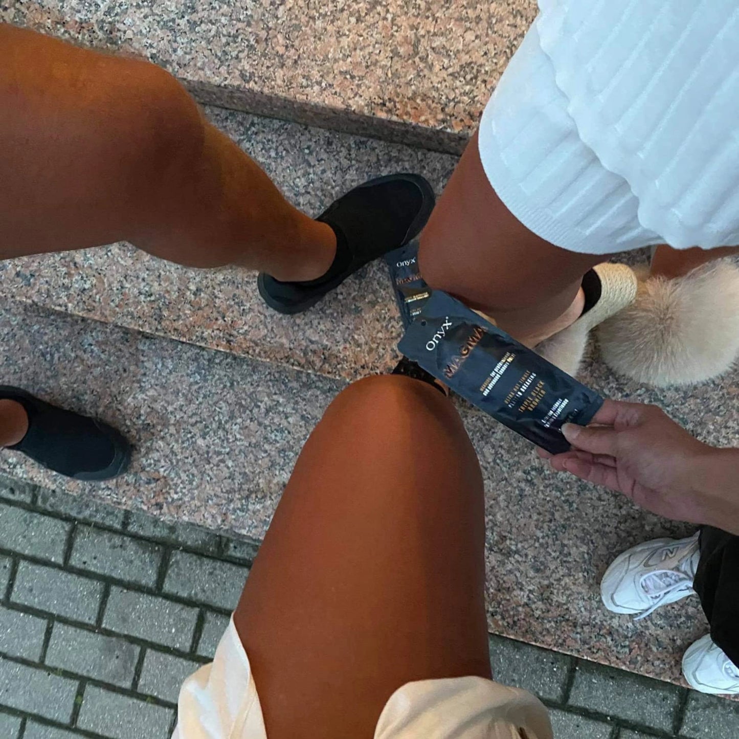 Loción Onyx Magma Tingle sostenida en la mano, con enfoque en las piernas bronceadas de varias personas en atuendo veraniego sobre escaleras de piedra