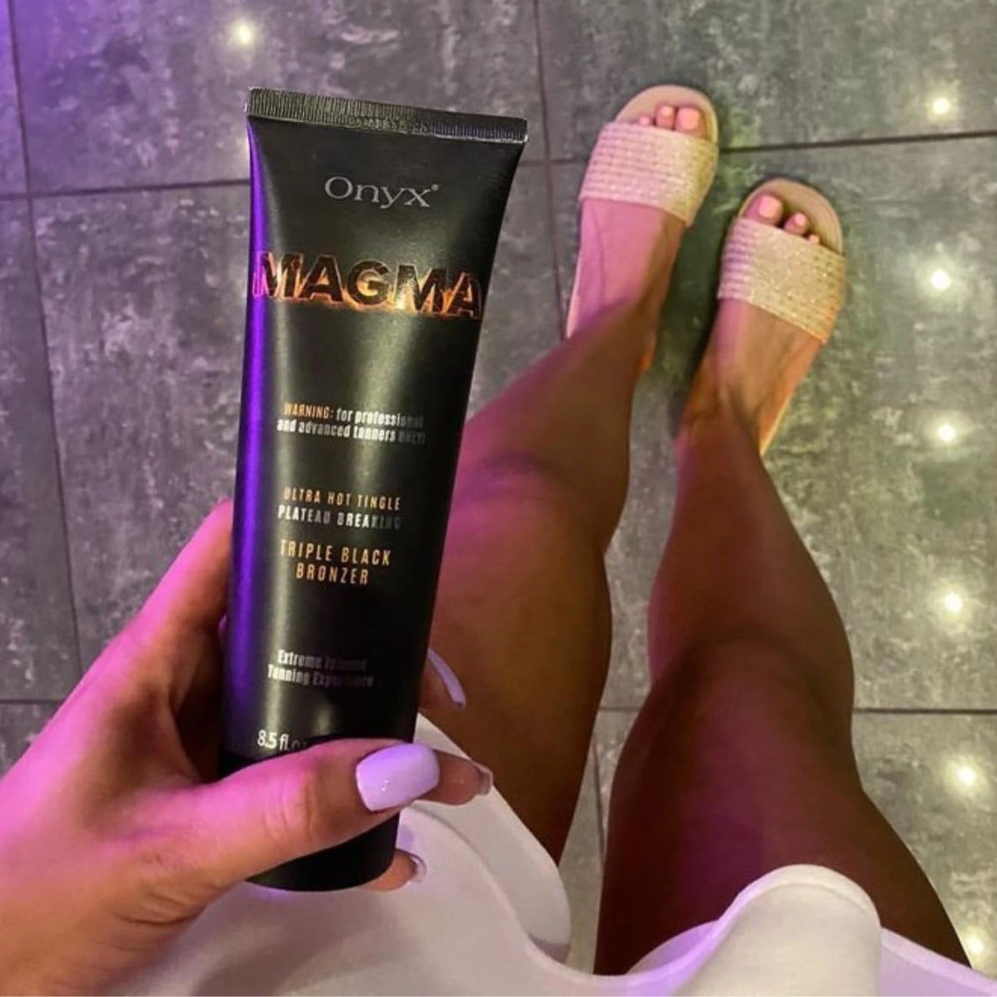 Primer plano de piernas bronceadas de una mujer sobre suelo brillante, sosteniendo el tubo Onyx Magma en la mano y destacando el bronceado intenso