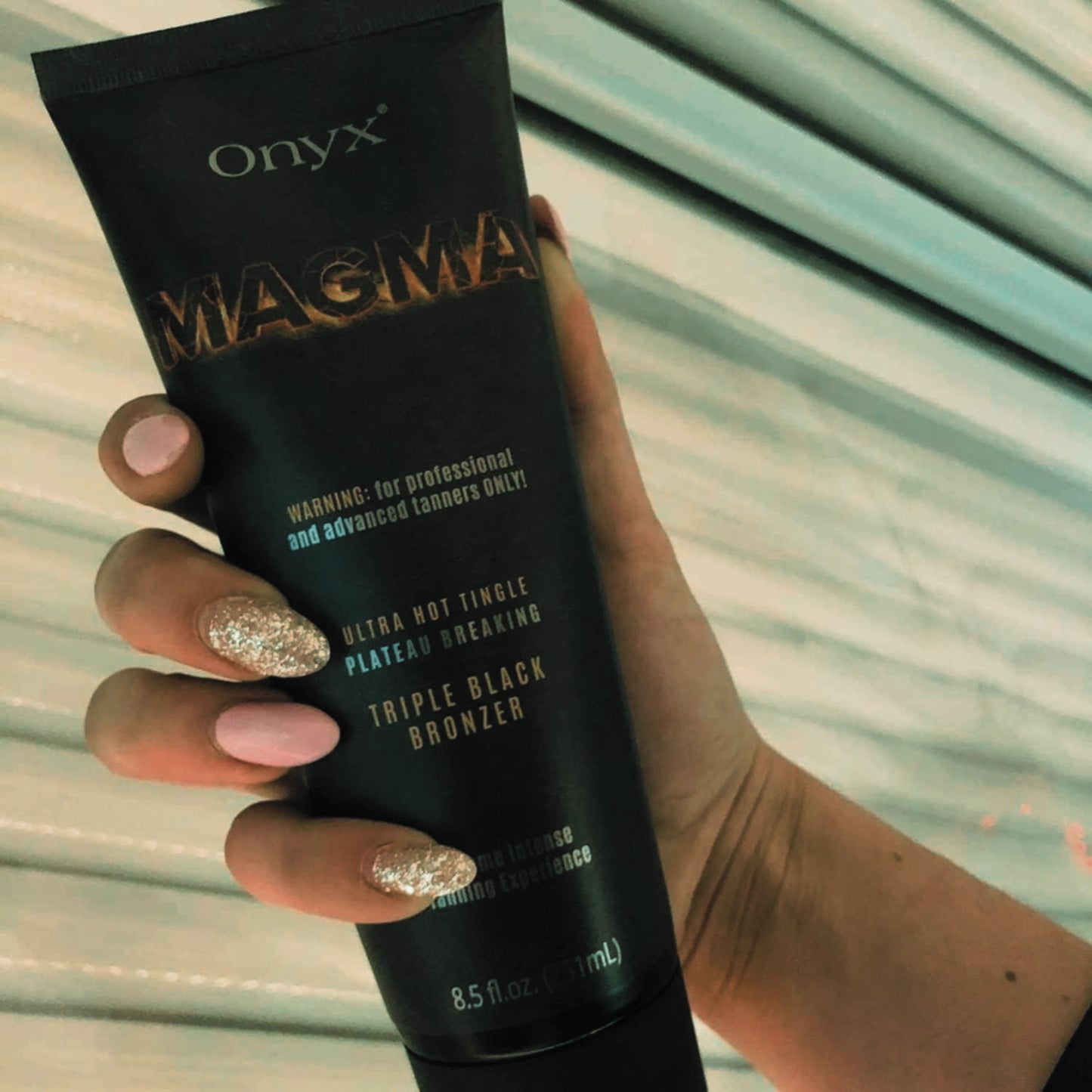 Mujer con diseño de uñas brillantes sostiene Onyx Magma Triple Black Bronzer frente a un solarium, mostrando un elegante detalle para bronceado indoor
