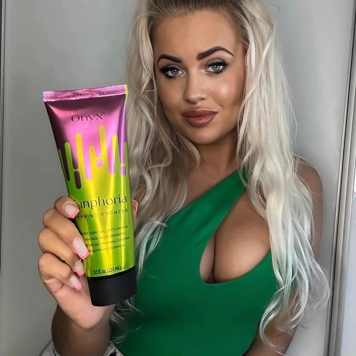 Mujer rubia con vestido verde sosteniendo un envase de Onyx Tanphoria Tanning Bronzer, mostrando el producto en primer plano