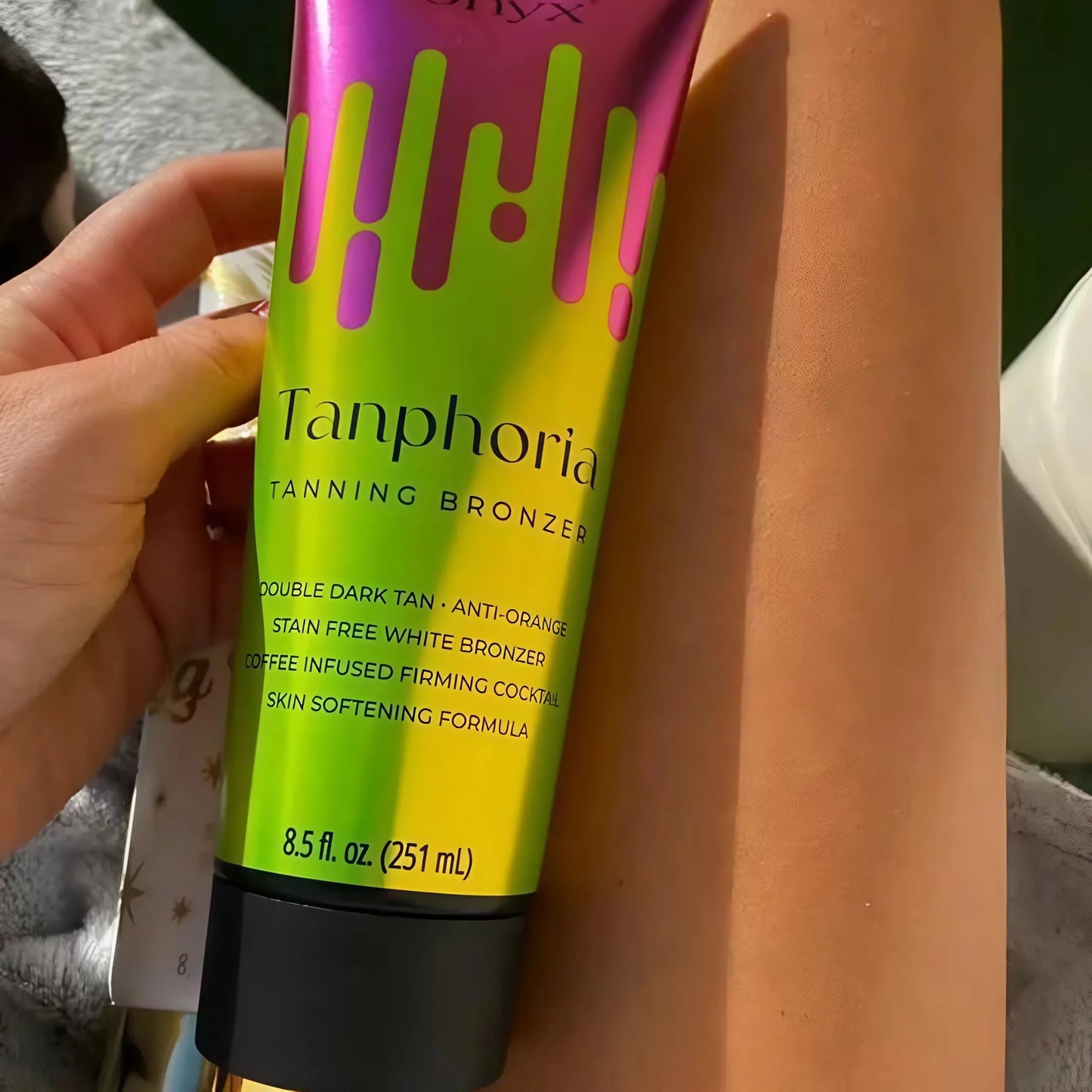 Envase de Onyx Tanphoria Tanning Bronzer sostenido sobre pierna bronceada al sol, mostrando detalles del producto en primer plano