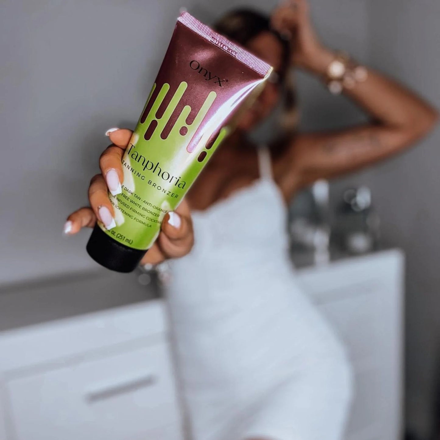 Primer plano del envase de Onyx Tanphoria Tanning Bronzer sostenido con la mano, fondo desenfocado de mujer con vestido blanco ajustado.
