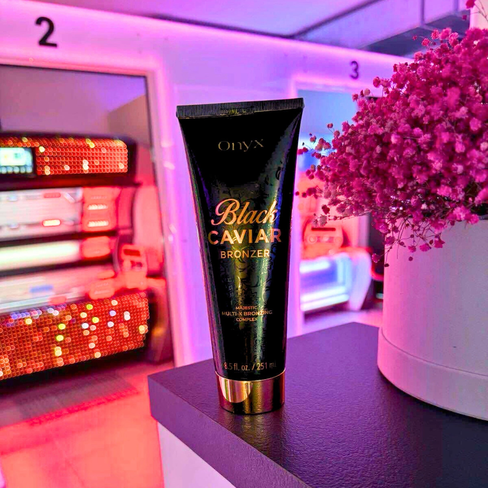 Loción bronceadora Onyx Black Caviar 251 ml colocada frente a un ramo de flores rosas y cabinas iluminadas en un estudio solar